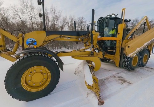 Equipment operator drives a road grader with snowplow attachment, clearing snow from a roadway in winter conditions.