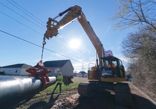 A construction worker guides a large pipeline as an excavator lifts it into place on a worksite near residential homes.