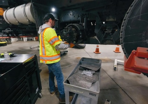 In a maintenance garage, a fleet maintenance technician works on the wheel assembly of a large truck while wearing high-visibility gear.