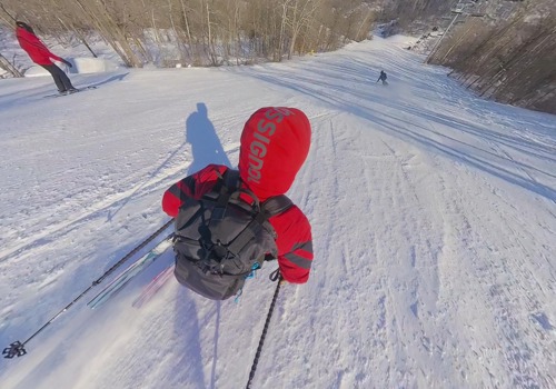 Skier in a red jacket and backpack glides downhill on a snowy slope at a ski resort, with other skiers visible in the distance.